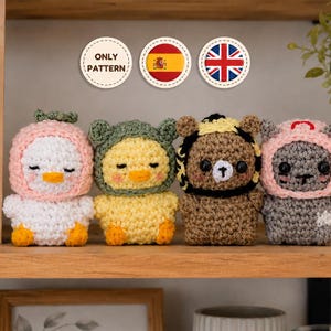 Puede incluir: Cuatro pequeños peluches de ganchillo hechos a mano dispuestos en fila. El primero es un pato blanco con capucha rosa, el segundo un pato amarillo con capucha verde, el tercero un oso marrón con capucha a rayas negras y amarillas, y el cuarto un gato gris con capucha rosa.