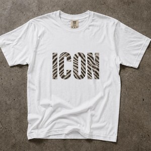 Könnte beinhalten: Ein weißes T-Shirt mit dem Wort "ICON" in einem Zebra-Print-Muster auf der Brust. Das T-Shirt besteht aus einem weichen Material und hat einen klassischen Rundhalsausschnitt. Das Kleidungsstück liegt flach auf einer strukturierten, grauen Oberfläche.