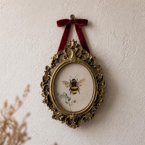 Puede incluir: Marco de fotos ovalado de color oro antiguo con una ilustración de abeja. El marco está adornado con detalles ornamentados y una cinta de terciopelo burdeos para colgar. La abeja es negra y amarilla.