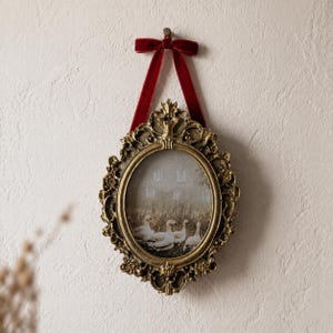 Puede incluir: Marco de fotos ovalado dorado con un diseño floral detallado. Se suspende con una cinta de terciopelo rojo atada con un lazo. La foto interior muestra gansos en un campo. El marco está colgado en una pared blanca.