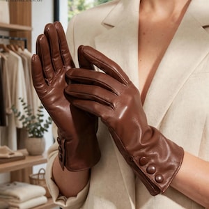 May include: A pair of smooth brown leather gloves featuring three matching circular buttons along the outer wrist cuff. The gloves have a classic tailored fit with visible stitching along the fingers and a soft, polished finish on the leather surface.