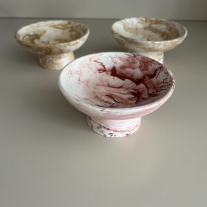 May include: Three small pedestal bowls with a marbled finish. One bowl features a white and deep red swirl pattern, while the two bowls positioned behind it display a white and tan marbled design. Each bowl has a wide, shallow basin and a sturdy base.