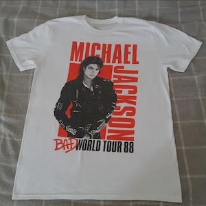 May include: A white short-sleeved t-shirt featuring a graphic print of Michael Jackson in a black leather jacket. The design includes bold red text reading MICHAEL JACKSON and black text reading BAD WORLD TOUR 88 on a red rectangular background.