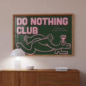 Puede incluir: Un cuadro rectangular enmarcado en madera con fondo verde oscuro y el texto DO NOTHING CLUB en letras rosas grandes. Un dibujo lineal estilizado en color rosa de una persona sosteniendo una copa de vino está centrado debajo del texto.