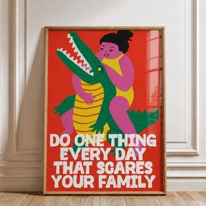 Puede incluir: Una l&aacute;mina rectangular enmarcada en madera con fondo rojo que muestra a una figura rosa en traje de ba&ntilde;o amarillo luchando con un cocodrilo verde de vientre amarillo. El texto blanco dice: DO ONE THING EVERY DAY THAT SCARES YOUR FAMILY.