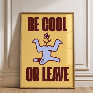 Puede incluir: Una lámina rectangular enmarcada con fondo amarillo y texto en rojo oscuro que dice BE COOL OR LEAVE. En el centro, una figura abstracta de color azul claro aparece representada boca abajo con una pequeña flor creciendo de su cuerpo.