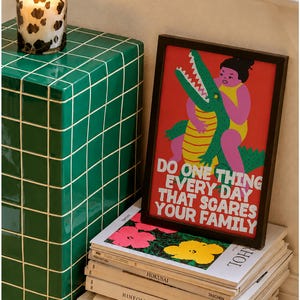 Puede incluir: Una impresi&oacute;n rectangular enmarcada con fondo rojo que presenta una ilustraci&oacute;n gr&aacute;fica de una persona en traje de ba&ntilde;o amarillo luchando con un cocodrilo verde. El texto dice: DO ONE THING EVERY DAY THAT SCARES YOUR FAMILY en letras blancas.
