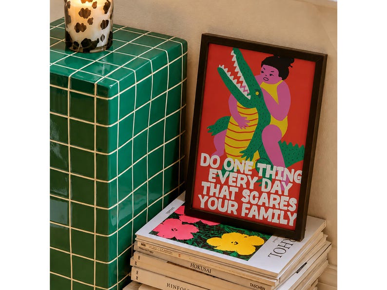 Puede incluir: Una impresi&oacute;n rectangular enmarcada con fondo rojo que presenta una ilustraci&oacute;n gr&aacute;fica de una persona en traje de ba&ntilde;o amarillo luchando con un cocodrilo verde. El texto dice: DO ONE THING EVERY DAY THAT SCARES YOUR FAMILY en letras blancas.