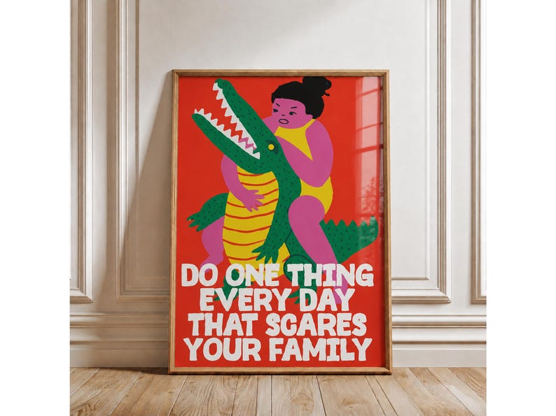 Puede incluir: Una l&aacute;mina rectangular enmarcada en madera con fondo rojo que muestra a una figura rosa en traje de ba&ntilde;o amarillo luchando con un cocodrilo verde de vientre amarillo. El texto blanco dice: DO ONE THING EVERY DAY THAT SCARES YOUR FAMILY.
