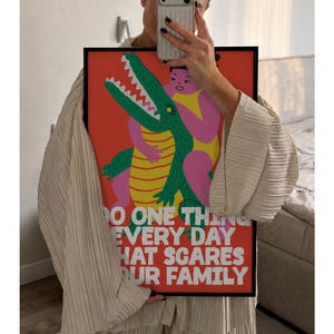 Puede incluir: Un p&oacute;ster rectangular enmarcado con un fondo rojo vibrante que presenta una ilustraci&oacute;n gr&aacute;fica de una figura rosa abrazando a un cocodrilo verde con vientre amarillo. El texto blanco dice: DO ONE THING EVERY DAY THAT SCARES YOUR FAMILY.