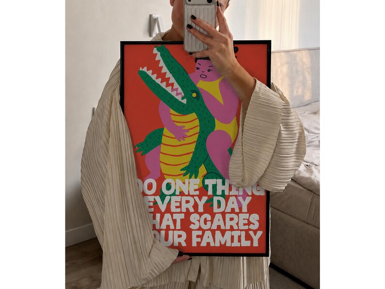 Puede incluir: Un p&oacute;ster rectangular enmarcado con un fondo rojo vibrante que presenta una ilustraci&oacute;n gr&aacute;fica de una figura rosa abrazando a un cocodrilo verde con vientre amarillo. El texto blanco dice: DO ONE THING EVERY DAY THAT SCARES YOUR FAMILY.