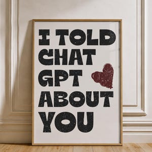 May include: A rectangular wall art print in a light wood frame featuring the black distressed text I TOLD CHAT GPT ABOUT YOU with a small, textured dark red heart graphic positioned to the right of the word CHAT on a white background.