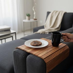Pode incluir: Uma bandeja flexível para apoio de braço feita de ripas de madeira conectadas sobre um sofá cinza escuro. Um prato de cerâmica branca com um biscoito redondo de gotas de chocolate e uma caneca de cerâmica preta estão sobre a bandeja.