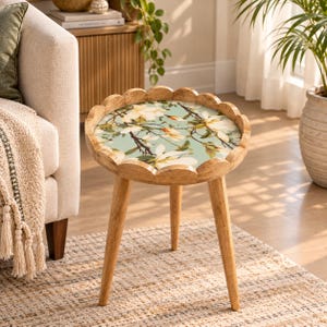 May include: A round wooden side table with three tapered legs and a scalloped edge. The tabletop features a light blue surface decorated with a detailed botanical print of white magnolia flowers and brown branches across the entire circular top.