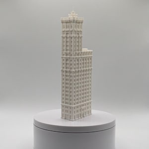 May include: A white 3D printed architectural model of a skyscraper with a stepped design, featuring multiple levels of windows and a central tower section, standing upright on a smooth, circular white base against a neutral background.
