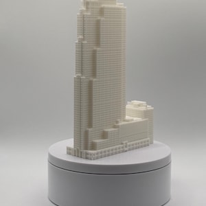 Puede incluir: Una maqueta arquitectónica blanca impresa en 3D de un rascacielos con diseño escalonado y una cuadrícula de pequeñas ventanas cuadradas, montada sobre una base circular blanca. La estructura presenta varios niveles y una torre central.