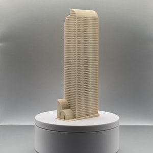 May include: A beige 3D printed architectural model of a tall skyscraper with a grid-like facade pattern and a smaller, lower building section attached to its base, featuring a curved roofline on the main tower and a flat rectangular base.