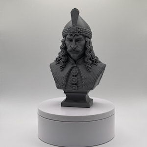 May include: A gray monochromatic bust sculpture depicting a figure with long wavy hair, a thick mustache, and a detailed helmet featuring a central crest. The figure wears a textured garment adorned with three circular emblems down the front.