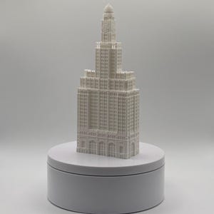 May include: A white 3D printed architectural model of a skyscraper with a tiered design, featuring a central tower, arched windows on the ground level, and a domed top, displayed on a plain white circular rotating display stand.
