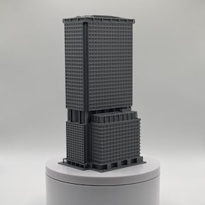 May include: A dark grey 3D printed architectural model of a skyscraper featuring a tall rectangular tower section connected to a shorter, wider base structure, both displaying a grid of small square windows across their exterior surfaces.