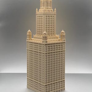 May include: A cream-coloured architectural model of a skyscraper featuring a rectangular base, a central tower section, and a domed roof. The structure displays a grid of numerous small windows and decorative corner turrets on the lower roof level.