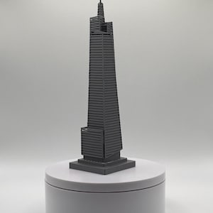 May include: A dark grey 3D printed architectural model of a skyscraper featuring a tapered, multi-tiered design with a central spire at the top. The structure stands on a square base and exhibits a detailed, ribbed exterior texture throughout.