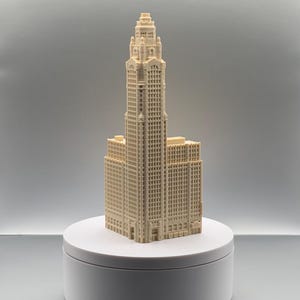 May include: A cream-colored 3D printed architectural model of a skyscraper featuring a central tower with a tiered top, numerous rectangular windows, and a multi-level base structure. The model is displayed on a plain white circular platform.