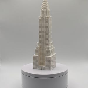 May include: A white 3D printed scale model of the Chrysler Building, featuring the iconic tiered skyscraper design with a pointed spire and detailed window grid patterns, standing upright on a circular white base against a plain background.