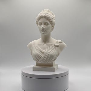 May include: A white matte bust sculpture of a person with wavy hair styled in an updo, wearing a draped garment with a circular brooch on the shoulder. The sculpture is mounted on a square base, which sits atop a cylindrical white display stand.