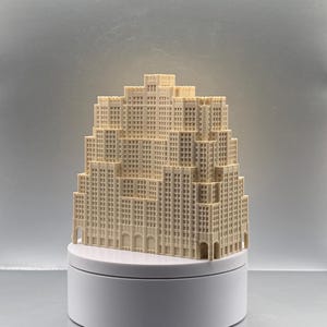 May include: A cream-colored 3D printed architectural model of a skyscraper with a stepped, tiered design. The structure features a grid of small square windows, vertical columns, and arched entryways at the base, all resting on a white circular stand.