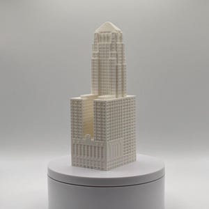 May include: A white 3D printed architectural model of a skyscraper featuring a tiered rectangular base and a central tower with a pointed roof. The structure displays a grid of small square windows across its facade, standing on a circular base.