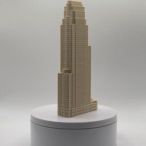 May include: A beige, three-dimensional architectural model of a skyscraper featuring a stepped, tiered design with a grid-like facade of windows. The structure is displayed on a plain, circular white base, showcasing vertical lines and levels.