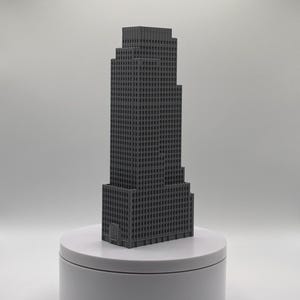 May include: A dark gray 3D printed architectural model of a skyscraper with a stepped design, featuring a grid of small square windows across its facade. The model is positioned upright on a smooth, circular, light gray display base.