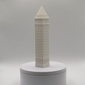 May include: A white 3D printed architectural model of a skyscraper featuring a rectangular base, numerous grid-patterned windows, and a pointed pyramidal roof. The structure stands upright on a smooth, circular white platform base.