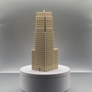 May include: A cream-coloured 3D printed architectural model of a skyscraper featuring a stepped design with multiple levels, a grid of small square windows, and a central spire at the top, standing upright on a circular white display base.