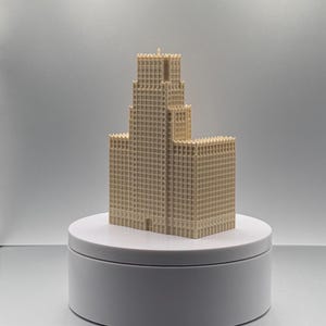 May include: A beige, three-dimensional architectural model of a skyscraper with a stepped, tiered design and a grid of small, rectangular windows covering the exterior facade, standing upright on a smooth, circular white display base.