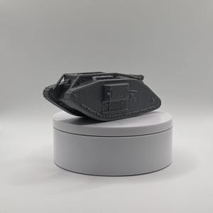 Pode incluir: Um modelo em escala cinza impresso em 3D de um tanque Mark IV da Primeira Guerra Mundial. A miniatura apresenta um casco em forma de romboide, lagartas proeminentes ao longo das laterais e sponsons laterais com pequenos canos de canhão.