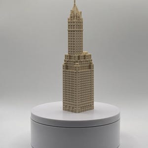 Könnte beinhalten: Ein beiges, dreidimensionales Architekturmodell eines Wolkenkratzers mit rechteckiger Basis, einem abgestuften Mittelteil und einer spitzen Turmspitze. Die Struktur zeigt ein Raster aus Fenstern und detaillierte Fassadentexturen.