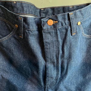 May include: Close-up of dark blue denim jeans. The jeans feature copper-colored rivets and a button closure. The pockets and seams are highlighted with contrasting stitching. The overall style is classic and durable.