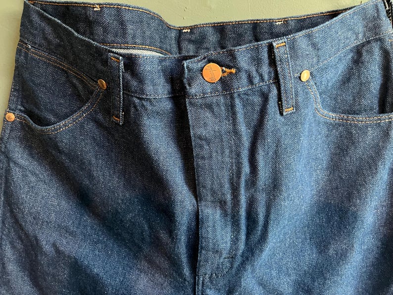 May include: Close-up of dark blue denim jeans. The jeans feature copper-colored rivets and a button closure. The pockets and seams are highlighted with contrasting stitching. The overall style is classic and durable.