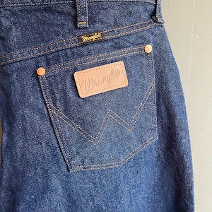 May include: Close-up of dark blue denim jeans featuring the Wrangler logo on the back pocket and waistband. The jeans have a classic five-pocket design with copper-colored rivets and stitching.