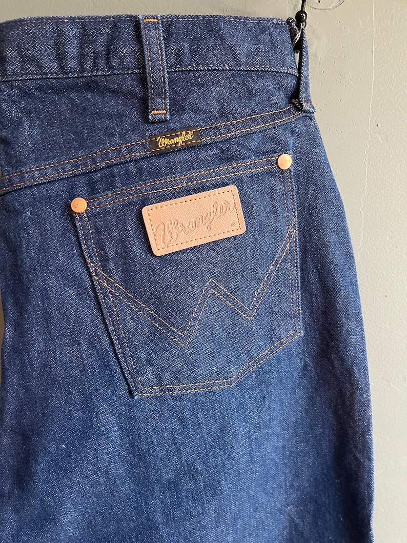 May include: Close-up of dark blue denim jeans featuring the Wrangler logo on the back pocket and waistband. The jeans have a classic five-pocket design with copper-colored rivets and stitching.