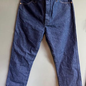 May include: Dark blue denim jeans with a button fly, five-pocket design, and copper rivets. The jeans are hanging against a neutral background, showcasing their classic style and construction. A timeless wardrobe staple.
