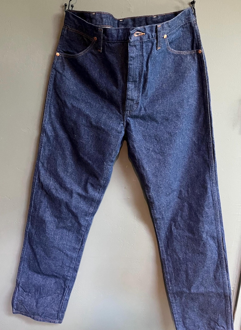 May include: Dark blue denim jeans with a button fly, five-pocket design, and copper rivets. The jeans are hanging against a neutral background, showcasing their classic style and construction. A timeless wardrobe staple.