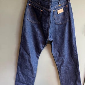 May include: Dark blue denim jeans with a classic five-pocket design. Features include copper rivets, a leather patch, and the Wrangler logo. The jeans are hanging on a wooden hanger.