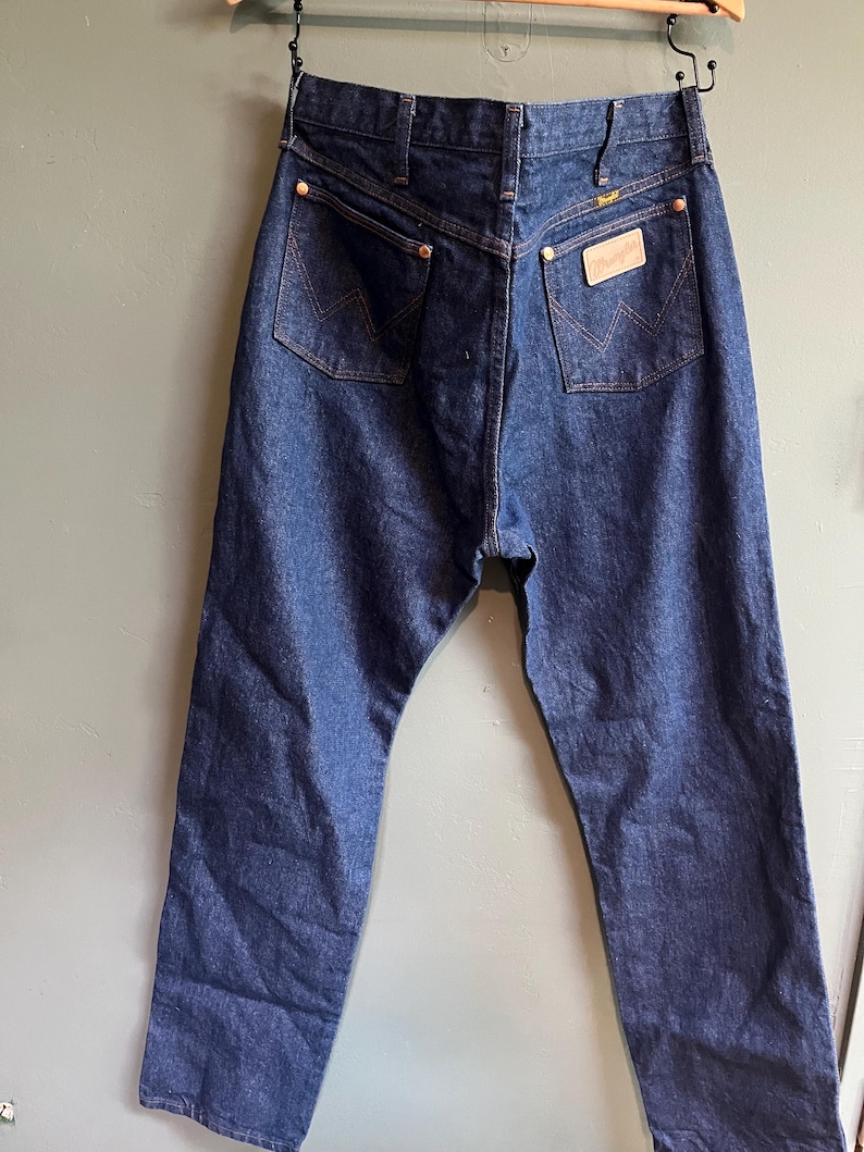 May include: Dark blue denim jeans with a classic five-pocket design. Features include copper rivets, a leather patch, and the Wrangler logo. The jeans are hanging on a wooden hanger.
