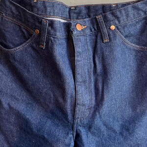 May include: Close-up of a pair of dark blue denim jeans. The jeans have a classic five-pocket design with copper-colored rivets and a button closure. The denim fabric has a slightly faded appearance.