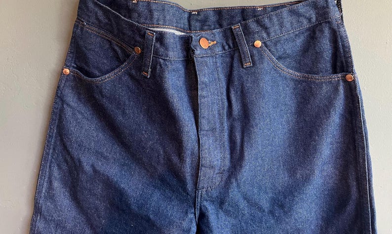 May include: Close-up of a pair of dark blue denim jeans. The jeans have a classic five-pocket design with copper-colored rivets and a button closure. The denim fabric has a slightly faded appearance.