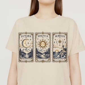 Puede incluir: Una camiseta beige con tres ilustraciones rectangulares estilo sello. El panel izquierdo muestra una luna creciente sobre montañas con el texto LUNARA DREAM BEYOND. El centro muestra un sol sobre un río con SOLIVIA CHASE THE LIGHT. A la derecha, un faro y una brújula con AURELIA GUIDE MY WAY.