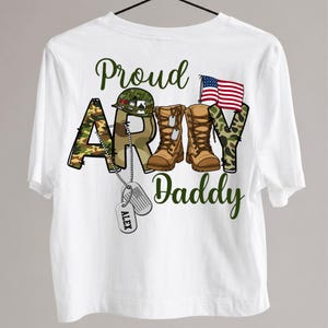 May include: White t-shirt with the words "Proud Army Daddy" in green script. The word "ARMY" is designed with camouflage print and military boots. An American flag and dog tags complete the design.